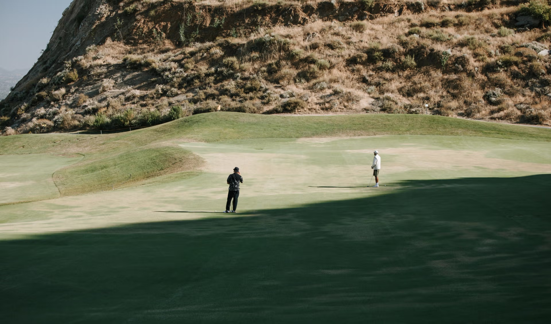 Two golfers on a green with rocky hillside backdrop